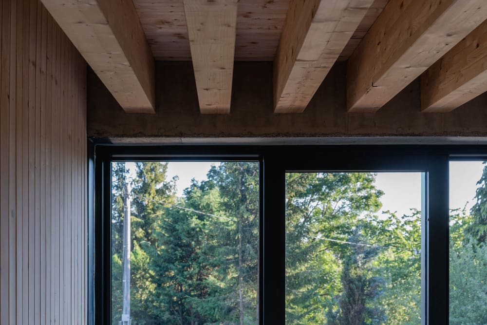 Wood-beamed ceiling meets a wood-paneled wall above black window frames, with green trees visible outside.