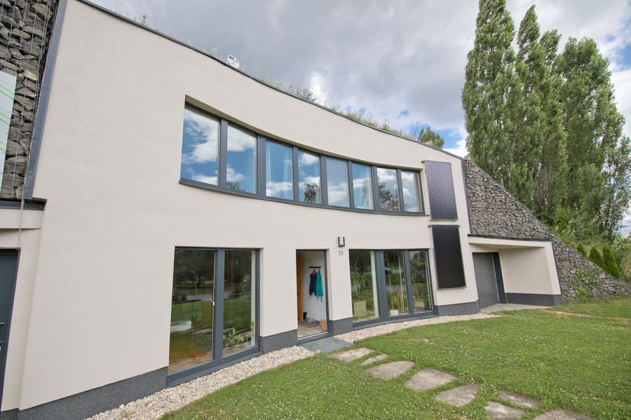 Zaoblená presklená fasáda podzemného domu, zatrávnená strecha, kamenný gabion a vchod s číslom 22